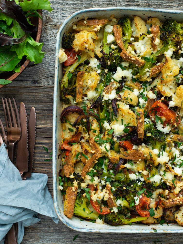 Bovenaanzicht: rechts ovenschotel broccoli en bloemkool met ei en feta op een houten tafel links bestek en een groene salade