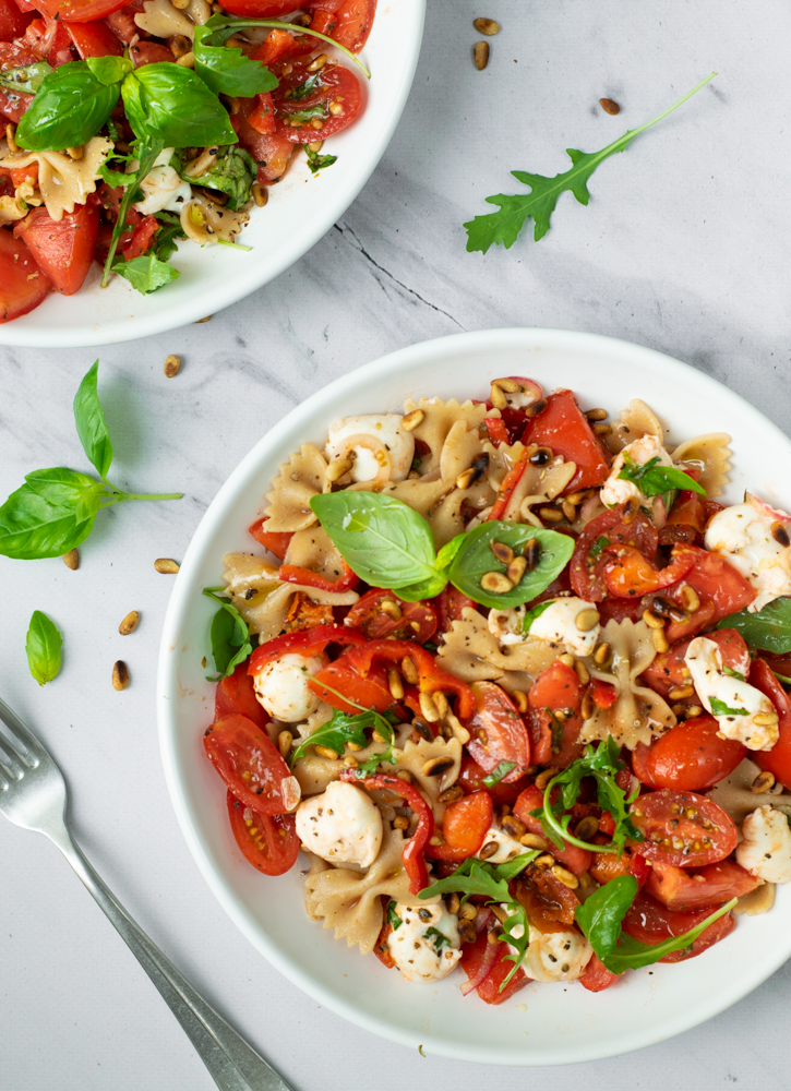 Een topshot van een wit bord gevuld met pastasalade caprese met farfalle, gesneden tomaten, kerstomaatjes, bolletjes mozzarella, verse basilicum en geroosterde pijnboompitjes.