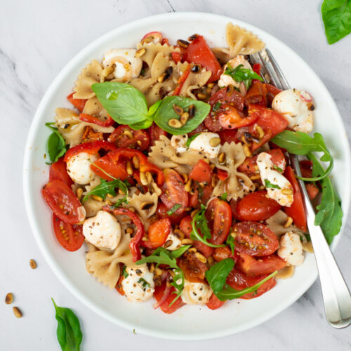 Een topshot van een wit bord gevuld met farfalle, gesneden tomaten, kerstomaatjes, bolletjes mozzarella, verse basilicum en geroosterde pijnboompitjes.