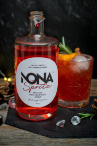 Een fles Nona Spritz met op de achtergrond een glas met de korlaalrode Christmas Spritz, afgewerkt met sinaasappelzeste en rozemarijn.