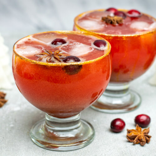 Twee bolvormige glazen gevuld met een oranjerode appel-cranberrymocktail, afgewerkt met gouden glitter.