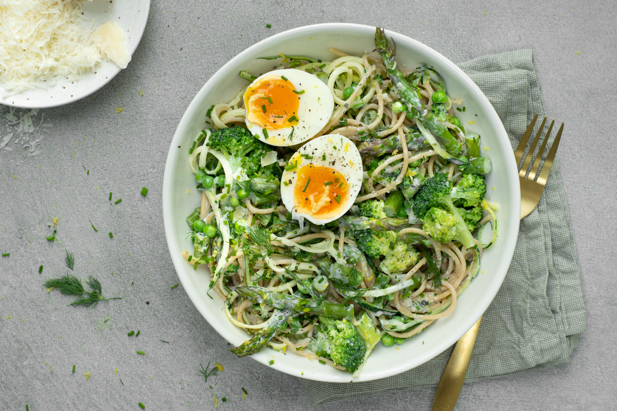 Bovenaanzicht van een spaghetti primavera met groene asperges, broccoli en erwtjes, afgewerkt met een gehalveerd zacht eitje.